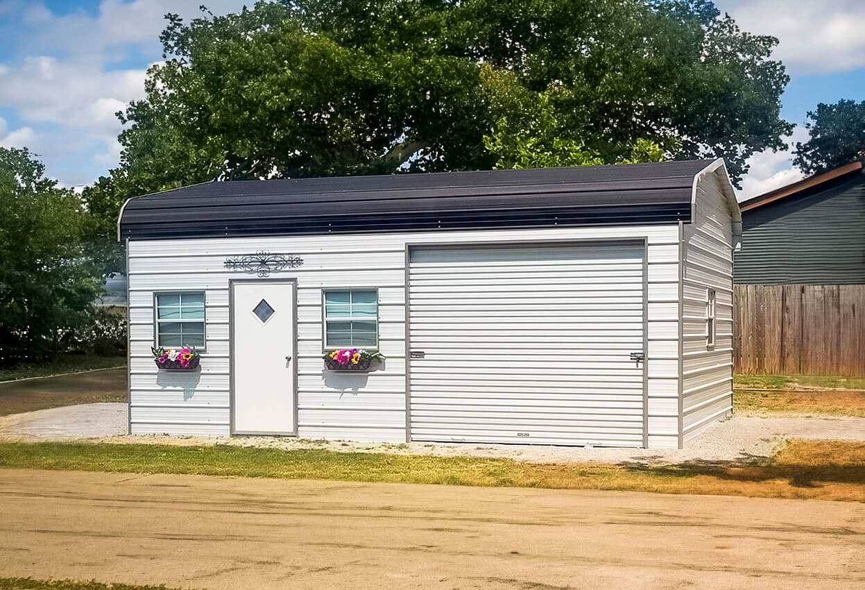 Small white metal barndominium with a curved black roof, featuring a white entry door with a diamond window, two windows with colorful flower boxes, a white roll-up garage door, and a decorative scrollwork accent above the door, set on a gravel pad with a large tree in the background.