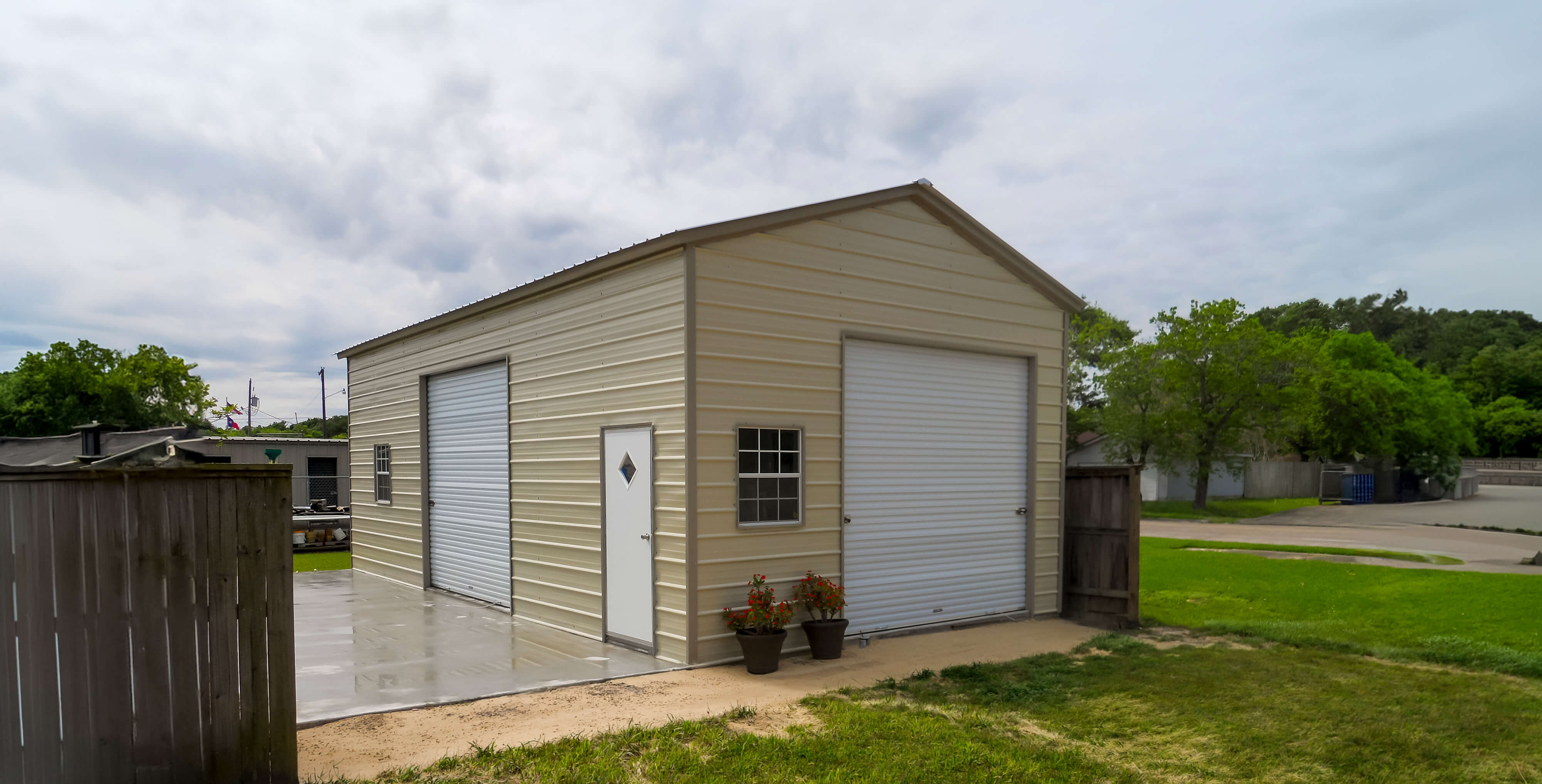 Beige metal shed house with two white roll-up garage doors, a side entry door, and windows.