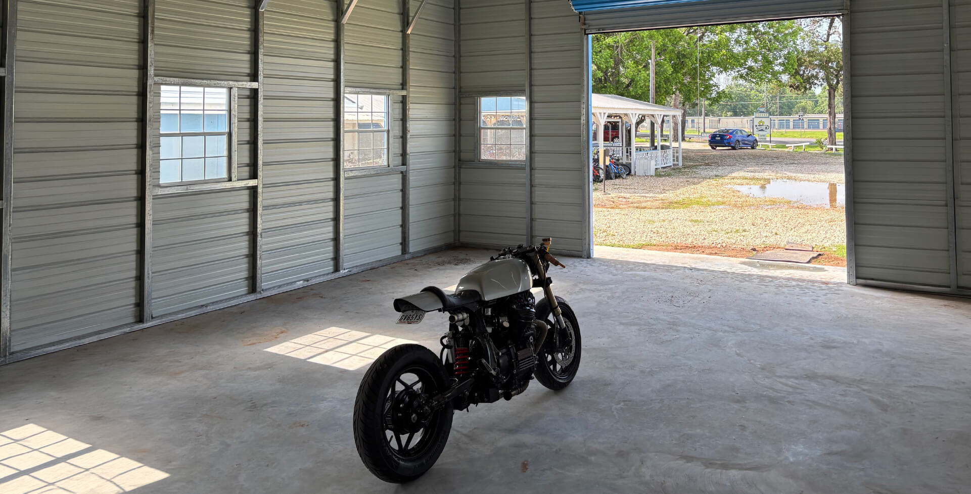 Motorcycle parked inside a spacious metal storage shed with windows, concrete flooring, and an open roll-up door.