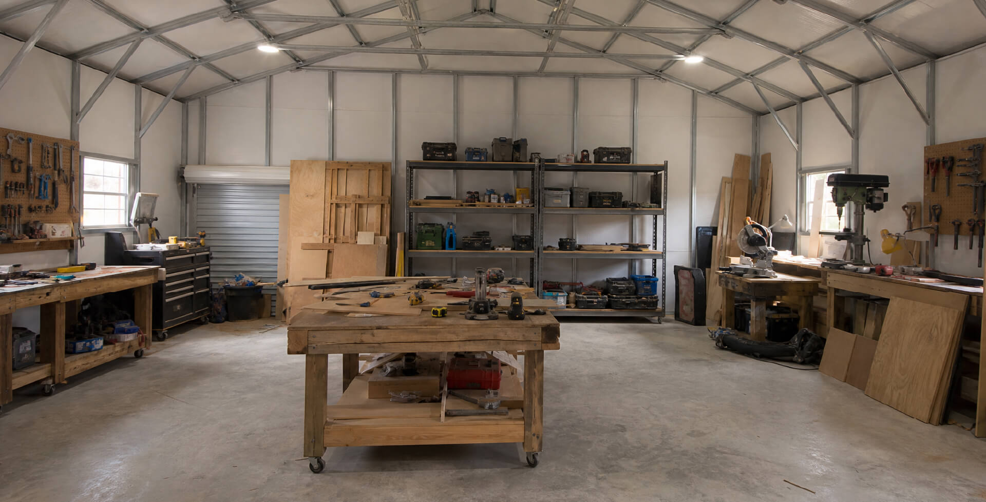 Spacious metal workshop with wooden workbenches, power tools, wall-mounted hand tools, shelving, storage, and concrete flooring.