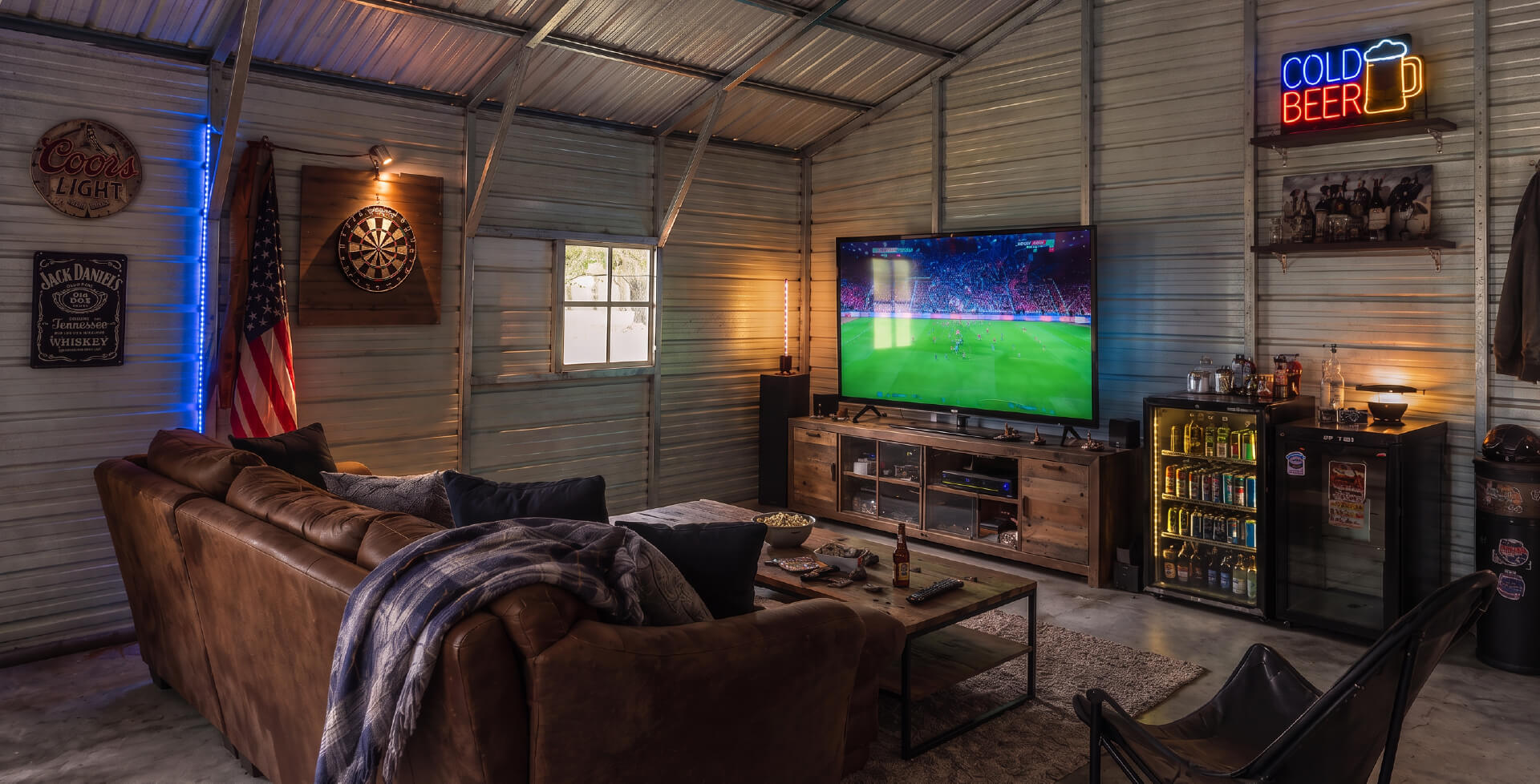 Finished metal building man cave with a sectional sofa, large TV, dartboard, beverage refrigerators, neon signs, and entertainment area.