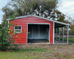 Red metal garage with white trim, open garage bay, window, and covered side porch surrounded by trees and grass.