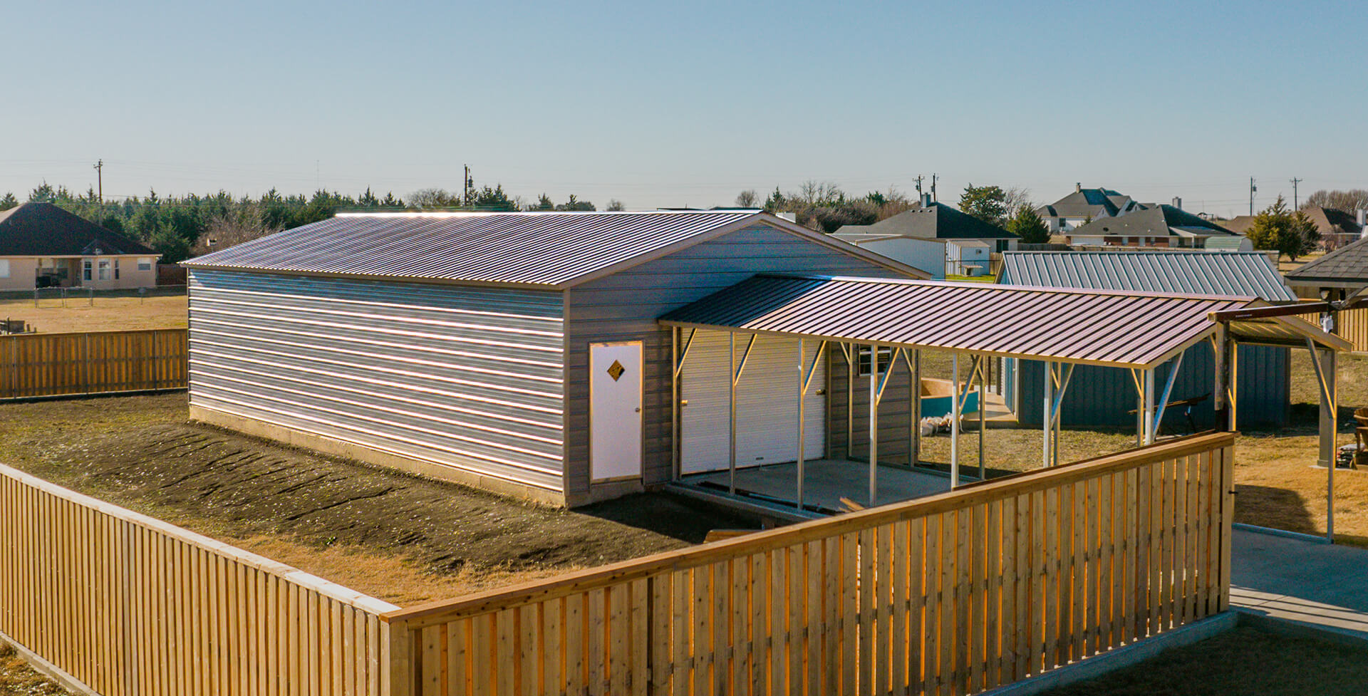 Gray metal garage building with a large roll-up door, walk-in entry door, and attached covered carport inside a fenced residential property.