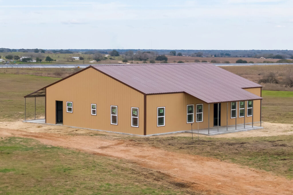 Large tan metal building with a brown vertical roof, multiple windows, covered entry areas, and open rural land surrounding the structure.