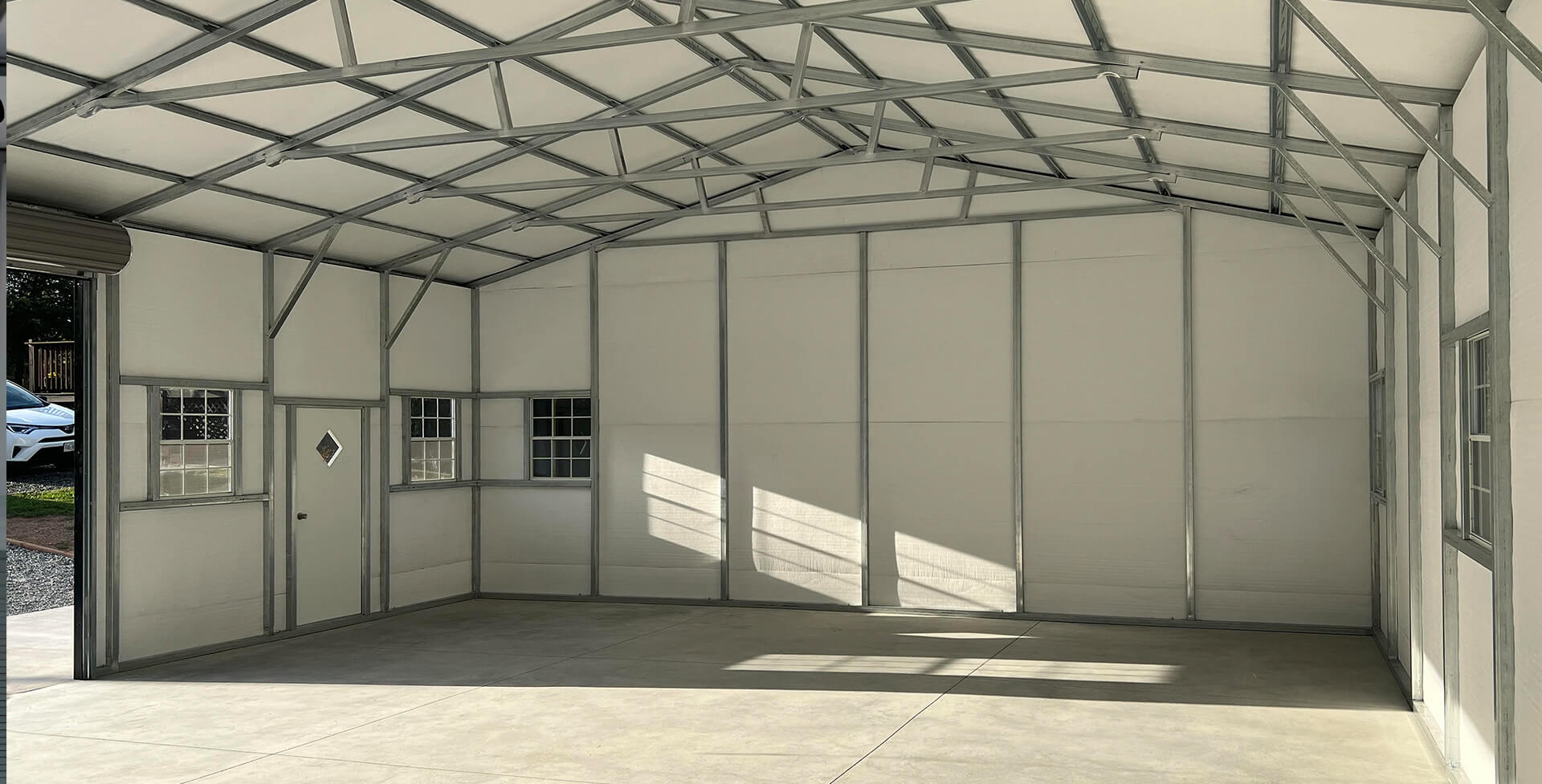 Interior of a metal building with insulated white wall and roof panels, exposed galvanized steel framing, windows, walk-in door, and concrete floor.