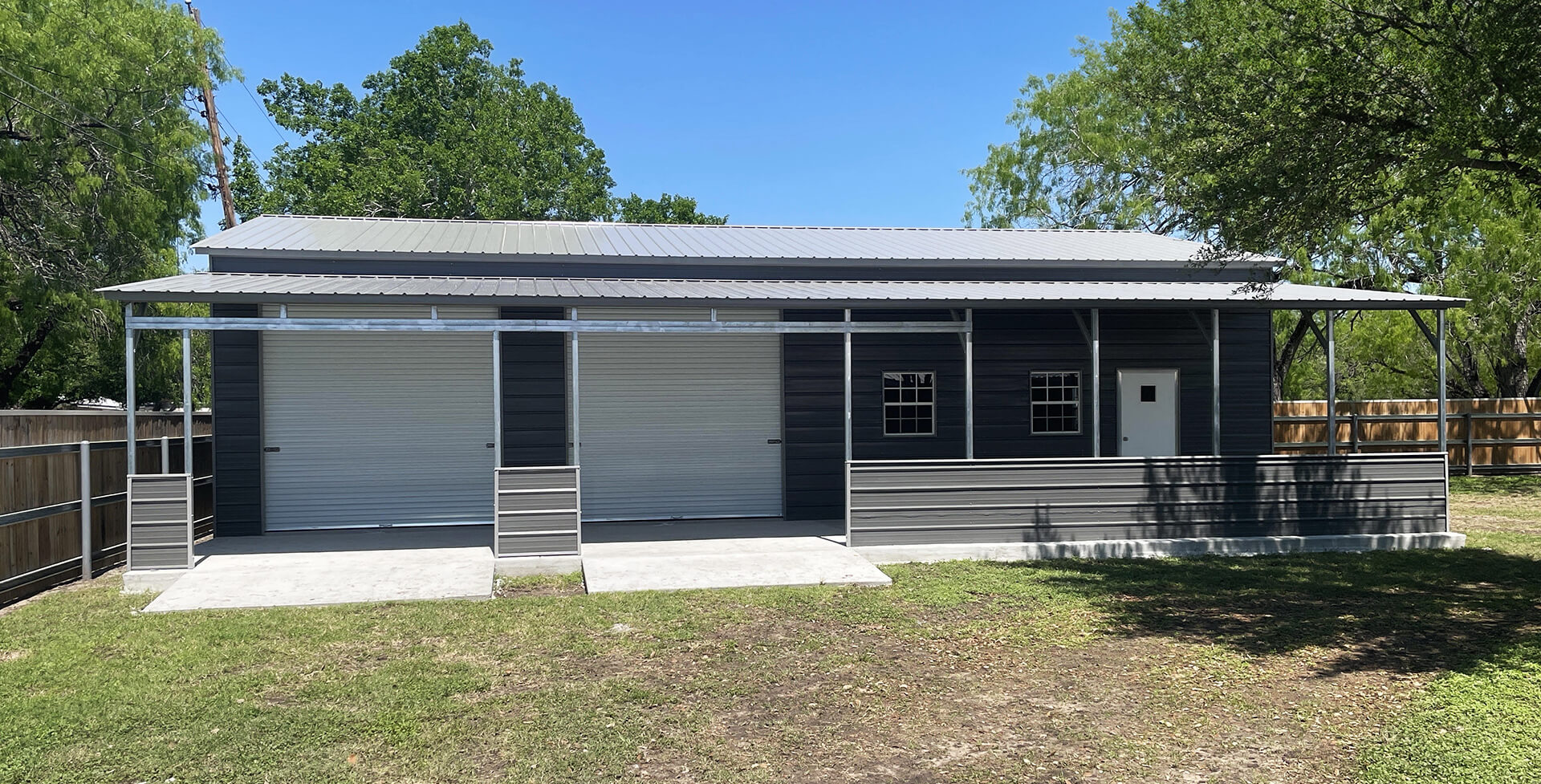 Large dark gray metal garage building with two roll-up garage doors, multiple windows, a walk-in door, and a covered front porch.